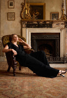 Woman in a black top and trousers  sitting in a chair in a room with a fireplace and decorative elements.