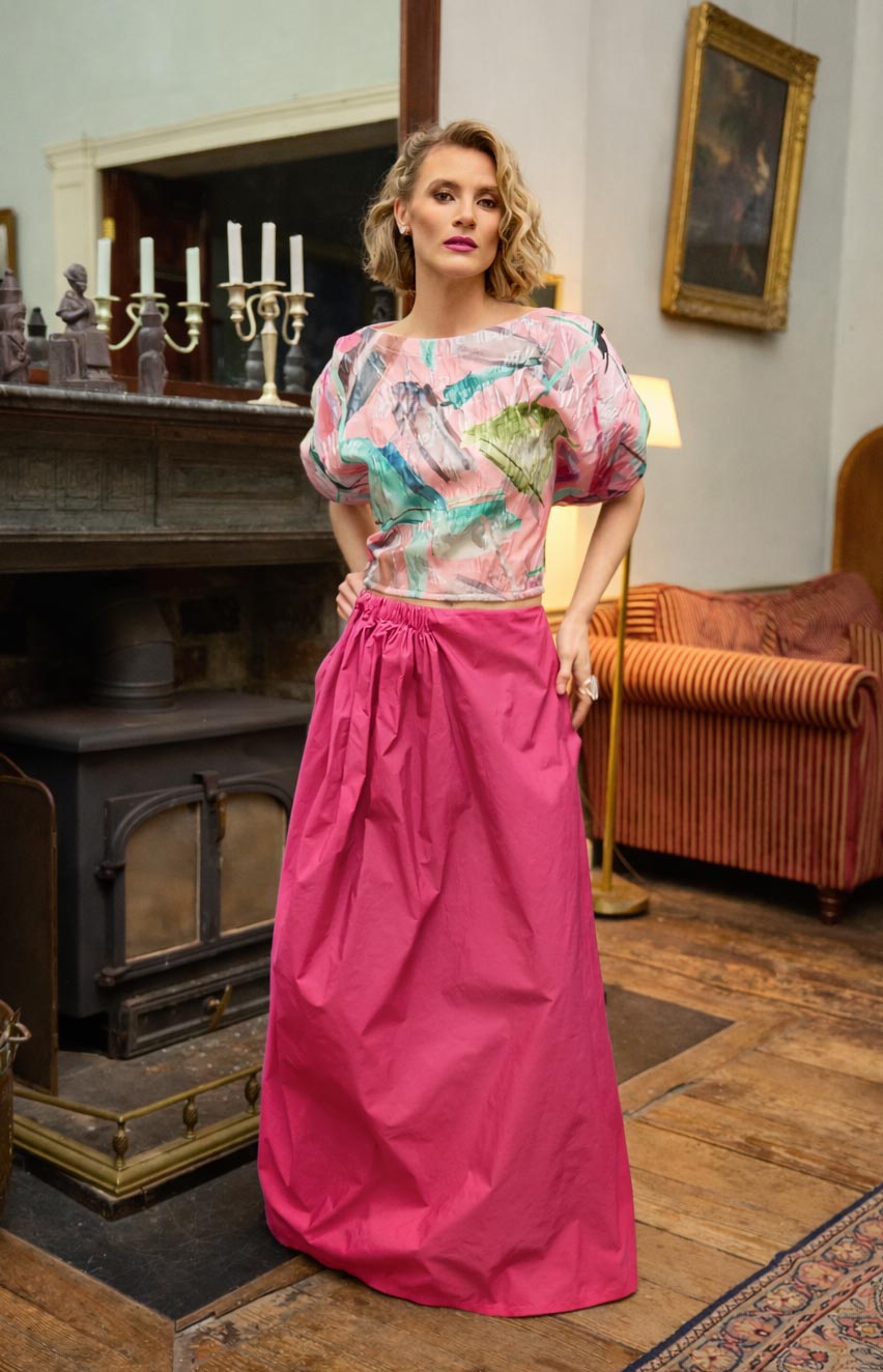 Woman in a colorful top and pink skirt standing in an elegant room with a fireplace and vintage decor.