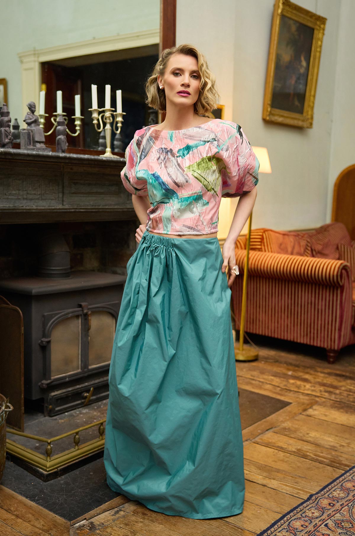 Woman in a colorful top and teal skirt standing in an elegant room with a fireplace and sofa.