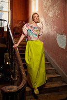 Woman in a colorful top and bright yellow skirt standing on a staircase with peeling paint walls.