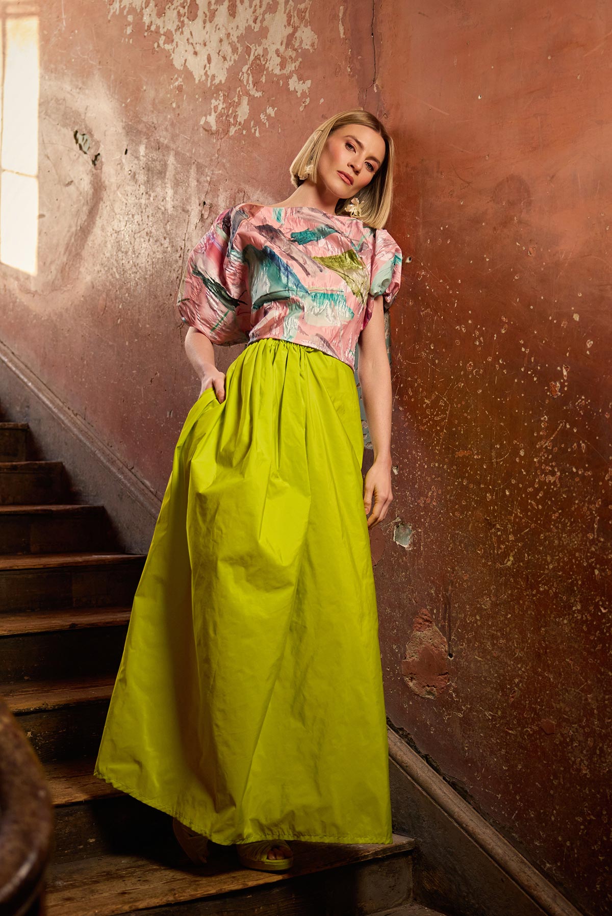 Woman in a colorful top and bright yellow skirt standing in an old, dilapidated building.