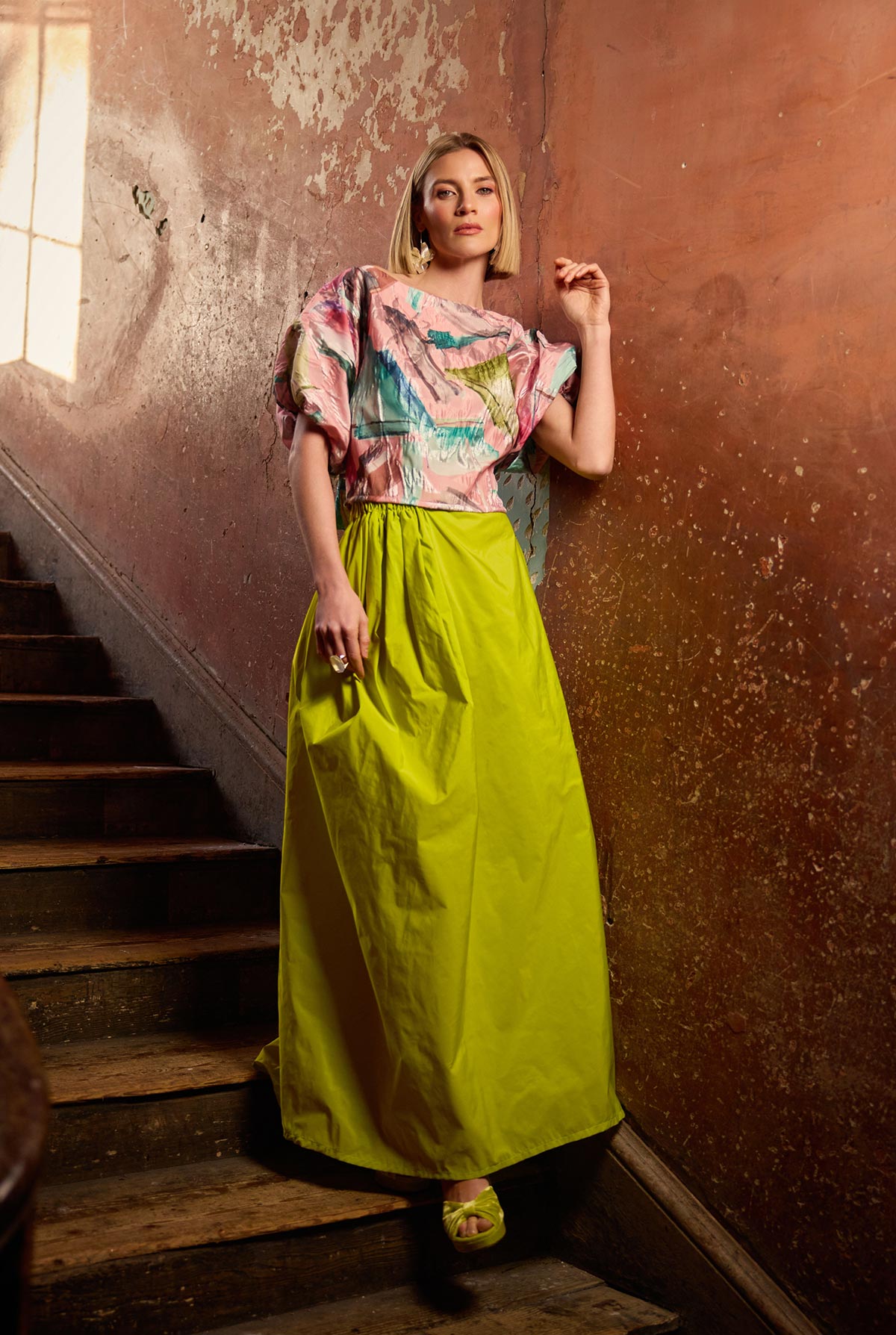 Woman in a colourful top and a yellow skirt standing on a staircase with a textured wall background.