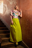 Woman in a colourful top and a yellow skirt standing on a staircase with a textured wall background.
