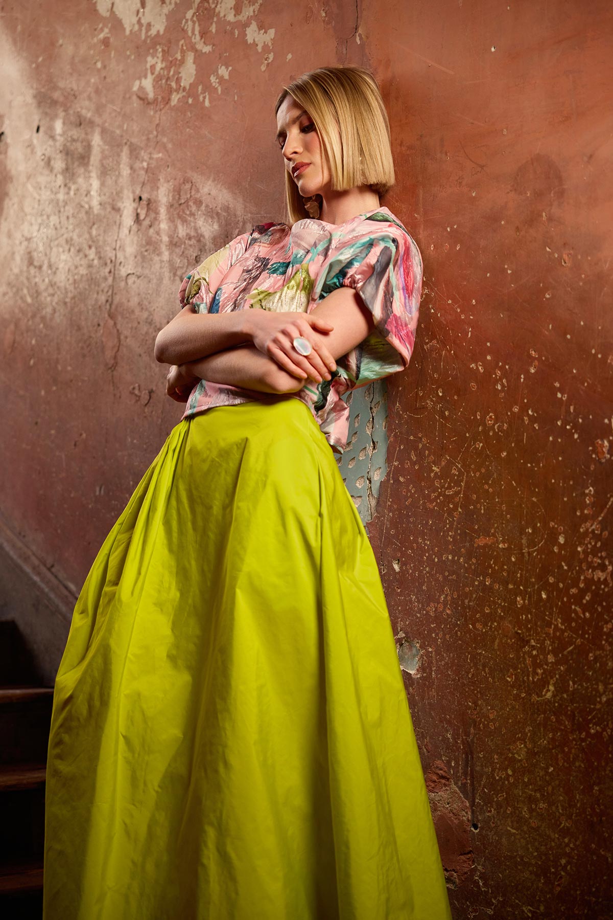 Woman wearing a colourful top and yellow skirt against a textured brown wall.