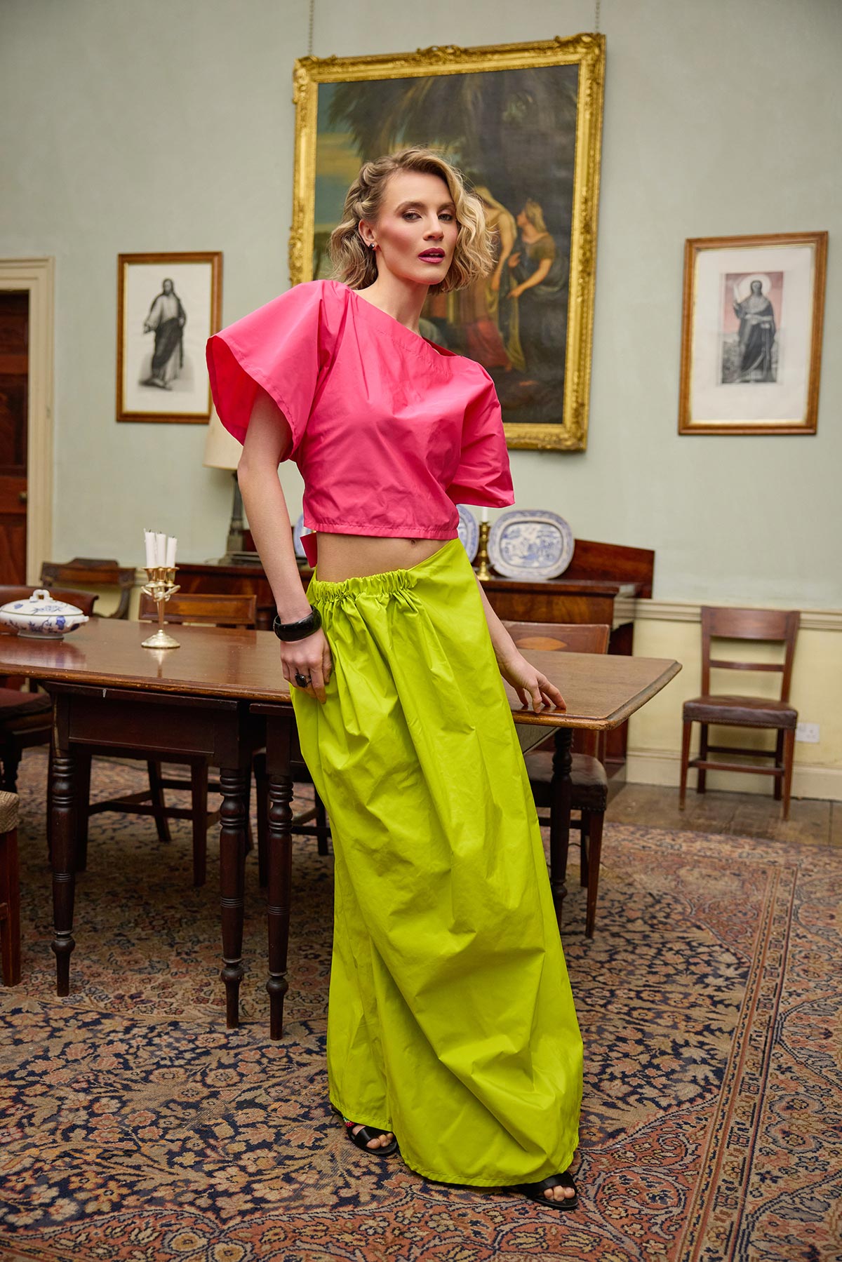 Woman in a pink top and lime yellow pants standing in an elegant room with wooden furniture and framed pictures on the wall.