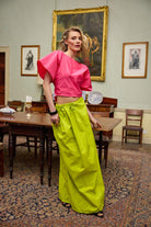 Woman in a pink top and lime yellow pants standing in an elegant room with wooden furniture and framed pictures on the wall.