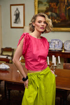 Woman in a pink top and lime yellow skirt standing in a room with a table and decorative items.