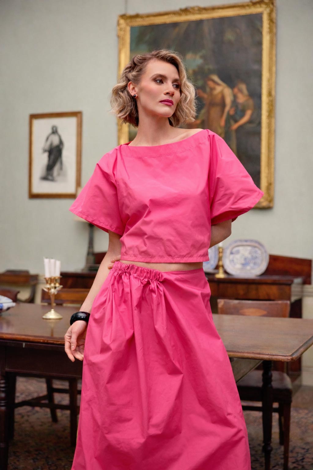 Woman in a bright pink outfit standing in an elegant room with paintings on the wall.