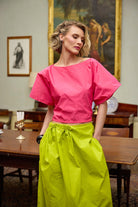 Woman wearing a bright pink top and lime yellow skirt in an indoor setting with a painting in the background.