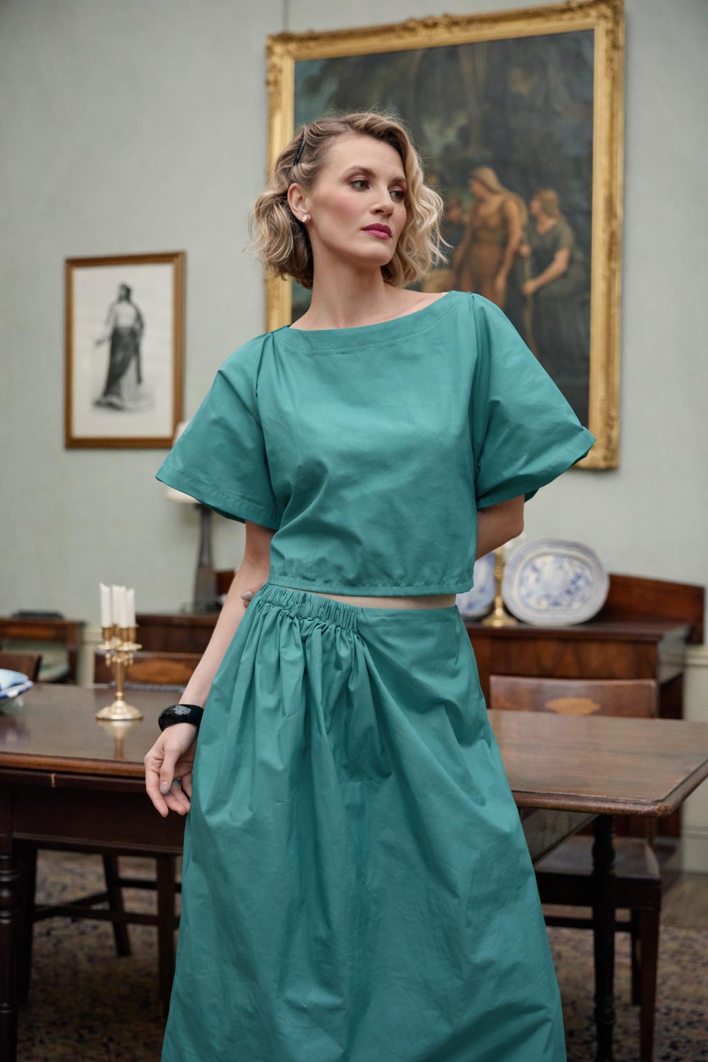 Woman wearing a teal outfit in an elegant room with paintings on the wall.