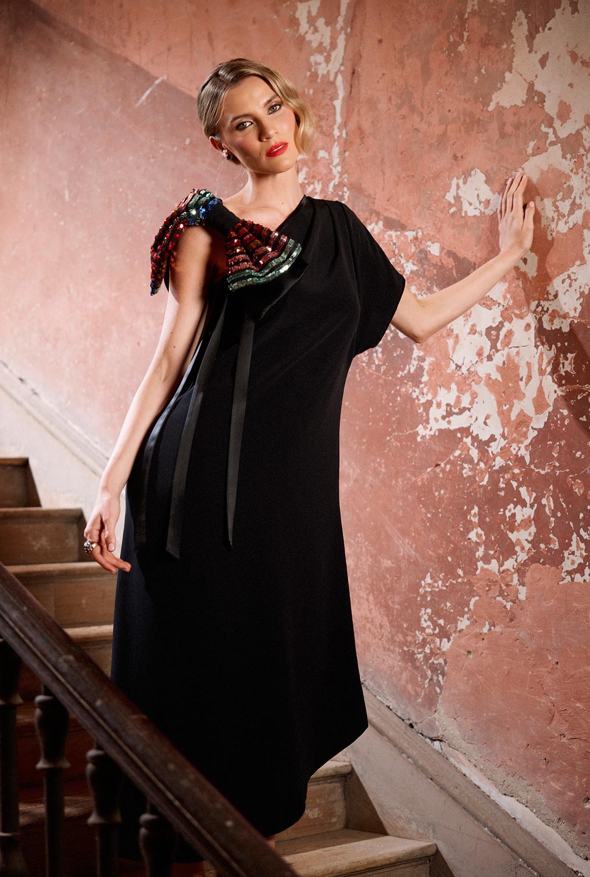Woman in a black dress with colorful shoulder bow standing on a staircase against a textured wall.
