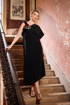 Woman in a black dress standing on a staircase with a pink wall and framed picture in the background