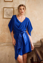 Woman wearing a blue dress with a belt standing against a beige wall.